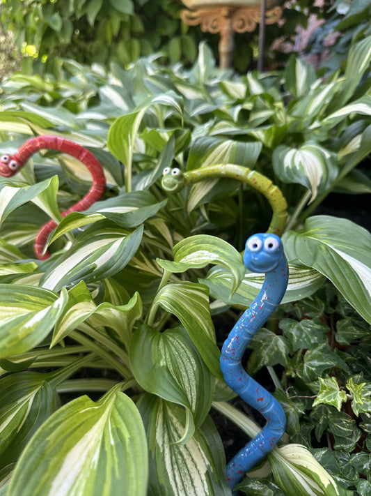 Close-up of red, blue, and green ceramic Wormyz garden stakes from JJ Potts Studio among garden foliage